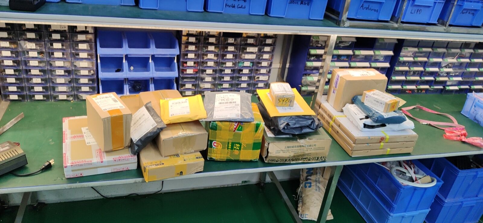 A shelf and bench lined with small parcels and blue parts bins, with samples ordered from several shops.